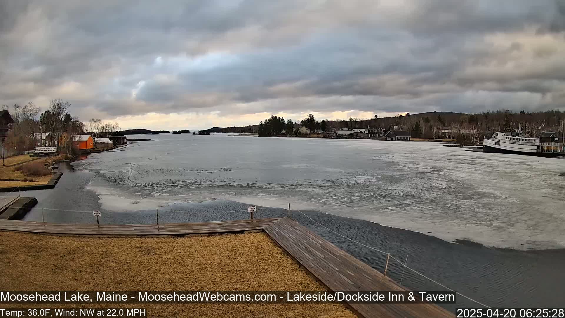 A partly frozen lake is viewed from a wooden dock under a cloudy sky, with buildings and boats visible in the distance.
