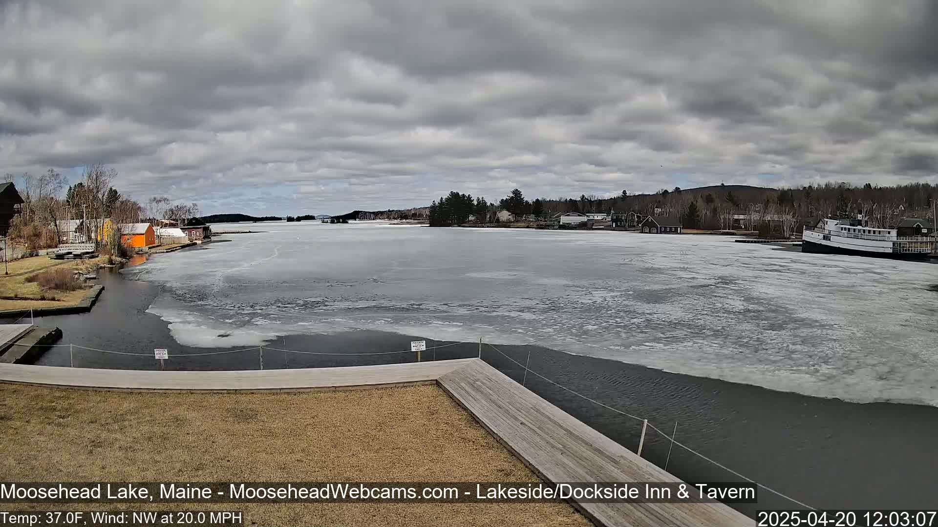 A partially frozen lake is seen from a wooden dock under a cloudy sky, with buildings and a boat visible in the distance.