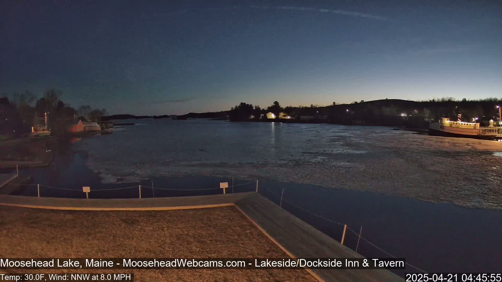 A twilight view of a partially frozen lake with a dock in the foreground, buildings on the far shore, and a boat moored near the shore under a clear, dark sky.