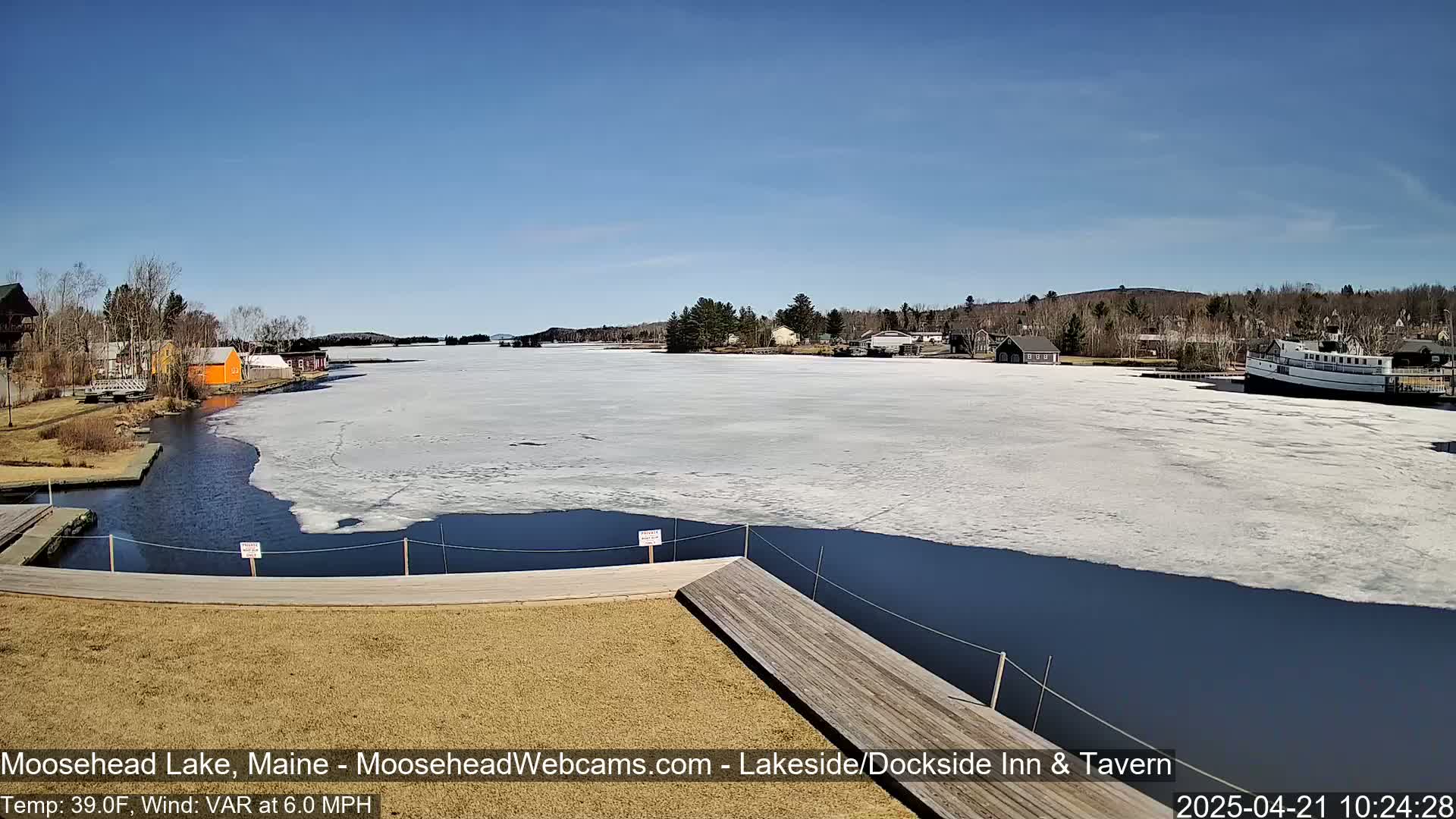 A partially frozen lake is viewed from a dock on a sunny day, with buildings and a boat visible in the distance.