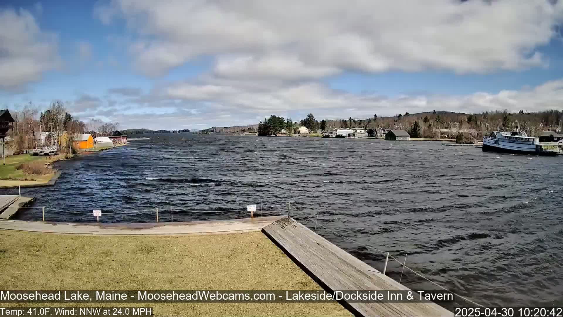A partly cloudy day with a 24 mph north-northwest wind shows choppy water on a lake with buildings and a boat visible in the distance.