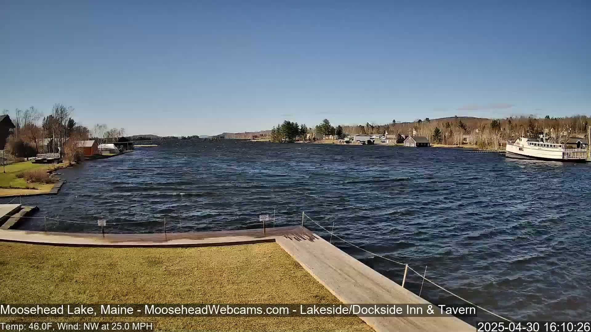 A lake with choppy water under a clear blue sky shows a docked boat and shoreline buildings in the distance.