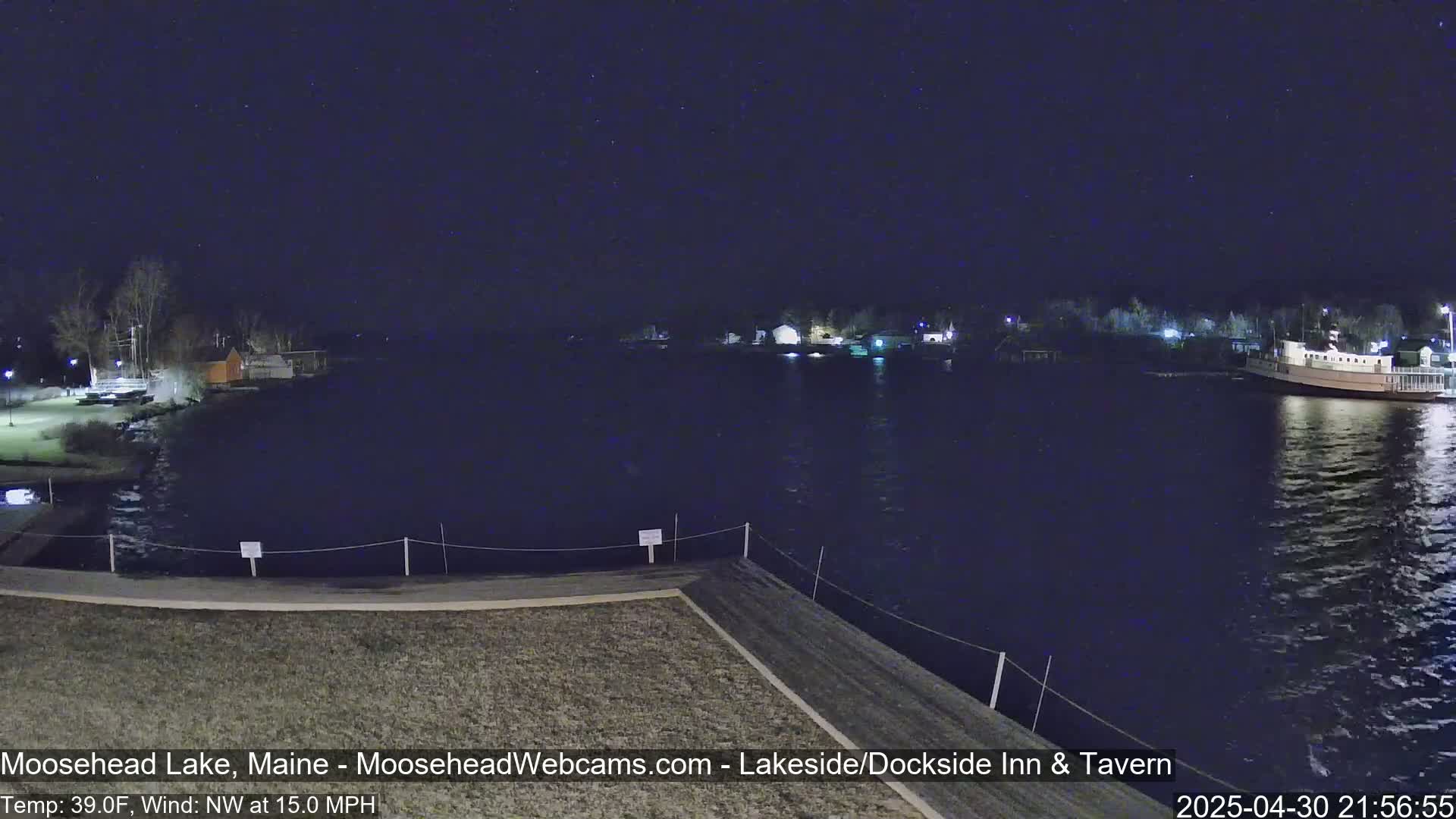 A nighttime view of a calm lake under a starry sky, with a shoreline featuring buildings and a docked boat, and a northwest wind blowing at 15 mph.