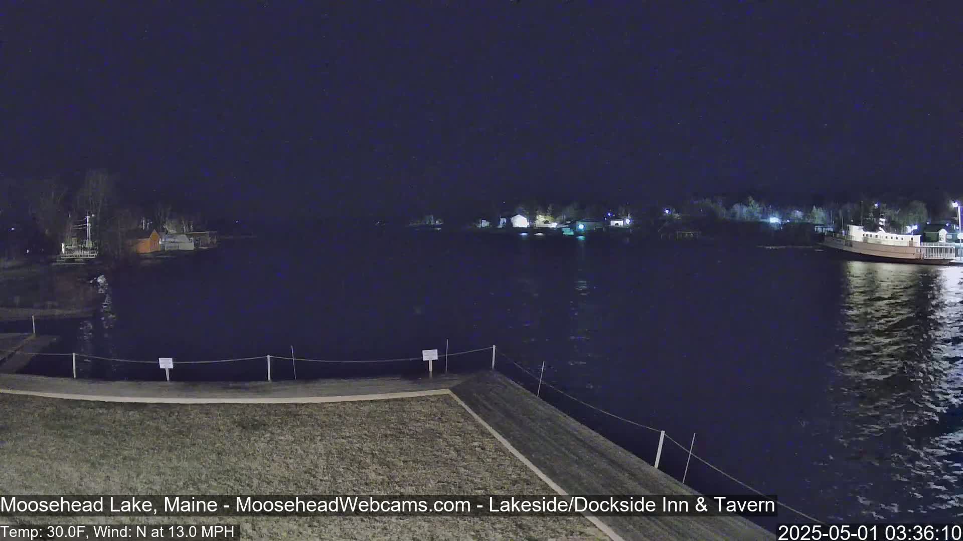 A dark, nighttime view of a lake with a boat docked and buildings along the shore, under a clear sky with a north wind of 13 mph and a temperature of 30°F.