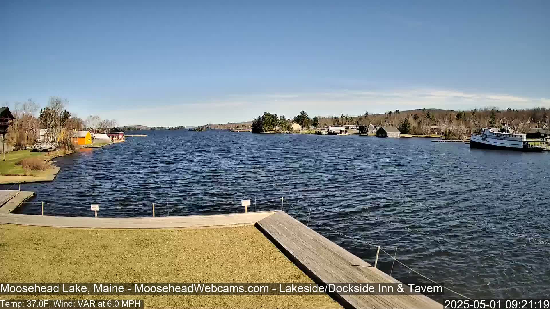 A sunny day on a lake shows a calm body of water with several buildings and boats along the shore, a wooden dock in the foreground, and a grassy area.