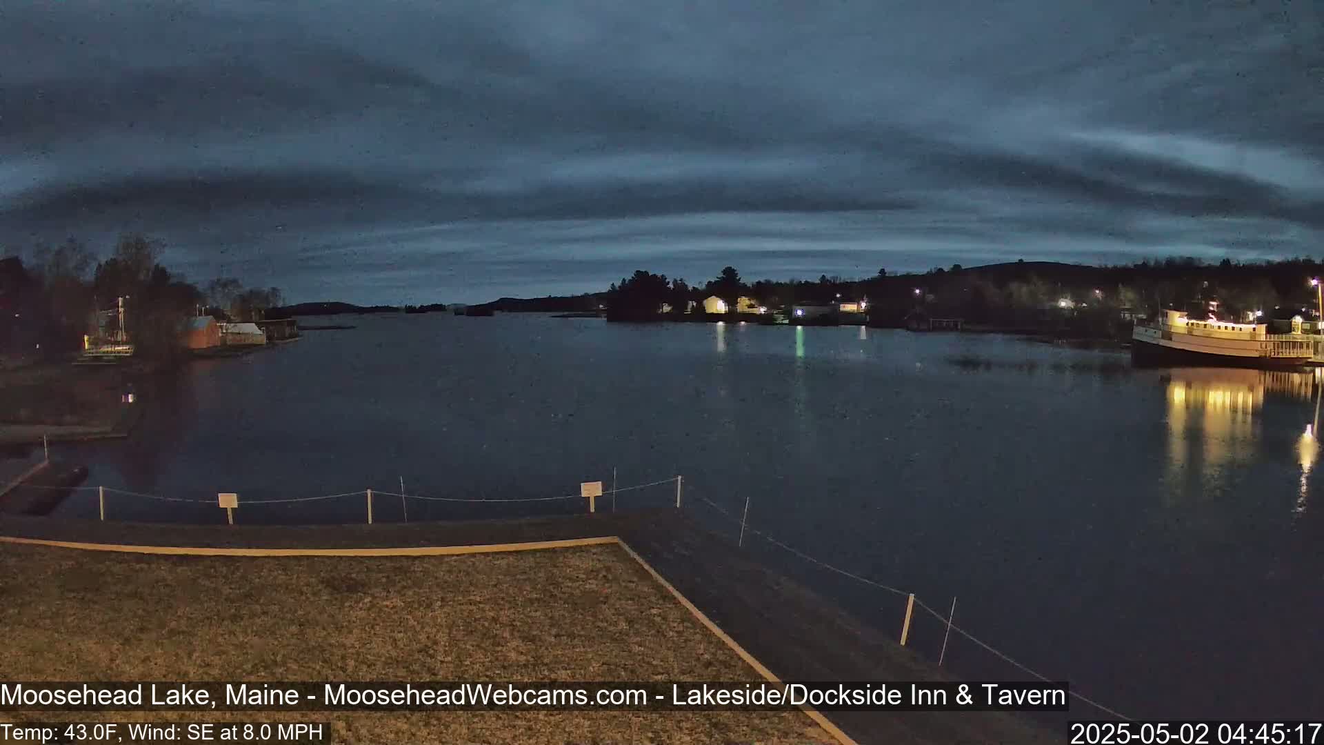 A dark, calm lake at night with a few lights visible on the shore under a cloudy sky, with a slight southeast wind.