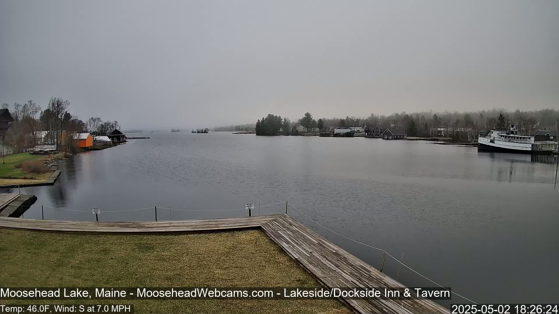 A gray, overcast day shows a calm lake with a wooden dock in the foreground, several buildings and boats along the shore, and trees lining the far bank.