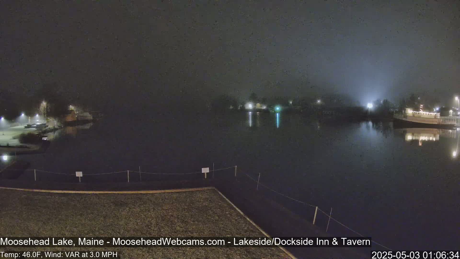 A nighttime view of a calm lake with fog, dimly lit buildings on the far shore, and a snow-covered area in the foreground.
