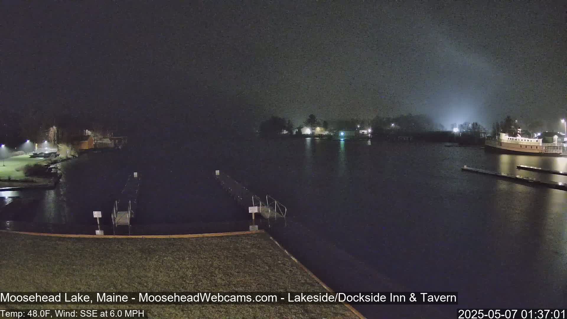 A nighttime view of a calm lake with docks, buildings, and a boat under a dark, possibly misty sky.