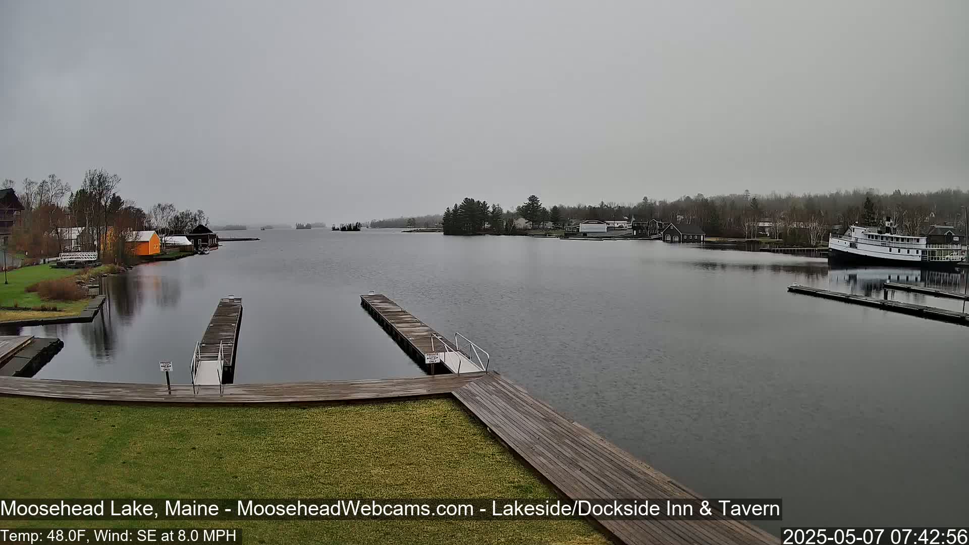 A lake view on an overcast day shows a calm body of water with docks, buildings, and a large boat moored at the far end.