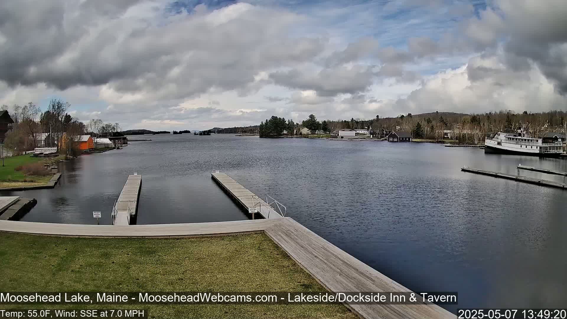 A partly cloudy day on a lake shows docks, buildings, boats, and a grassy area.