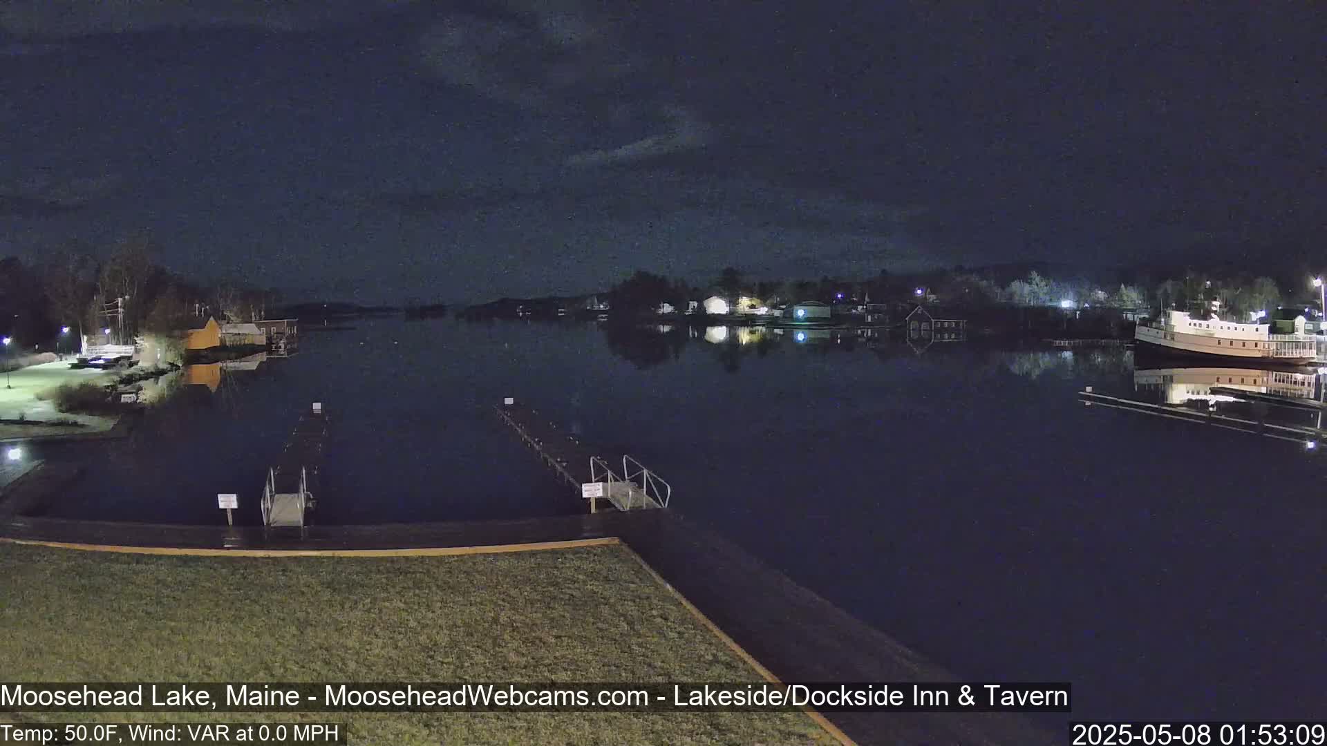 A nighttime view of a calm lake with docks and buildings visible on the shore under a dark sky.