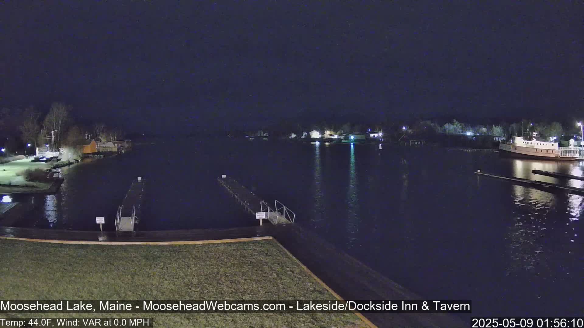 A nighttime view of a calm lake with docks, shoreline buildings, and a large boat, under a dark, clear sky.