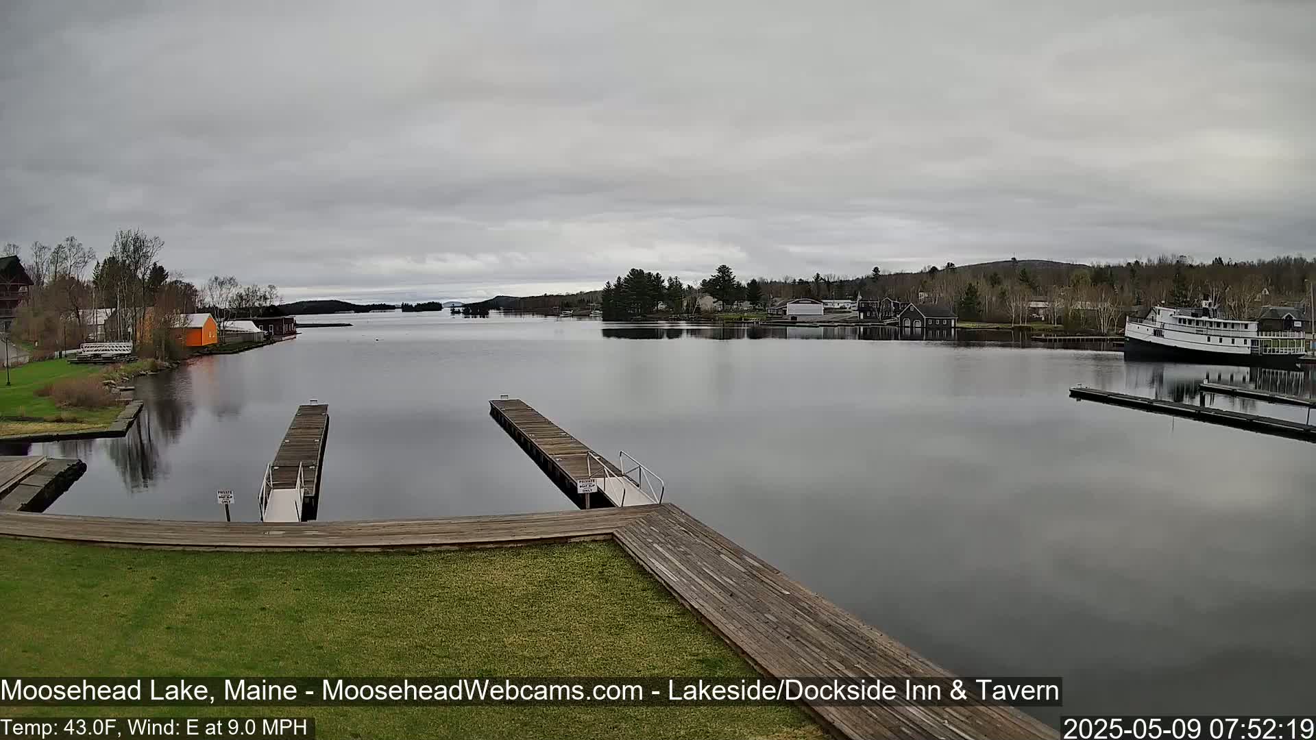 A calm lake with wooden docks, buildings along the shore, and a large boat under an overcast sky.
