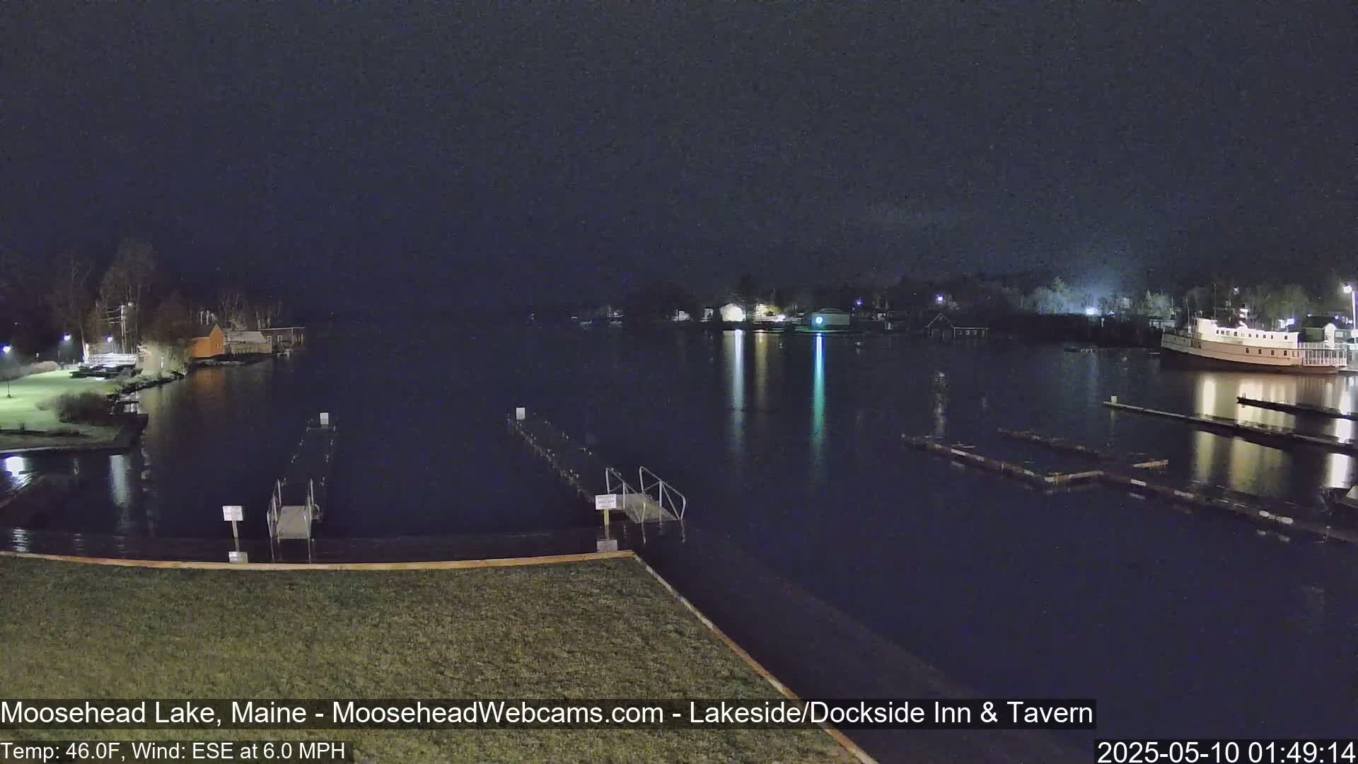 A dark nighttime view of a calm lake with docks, a boat, and buildings along the shoreline under a clear sky.