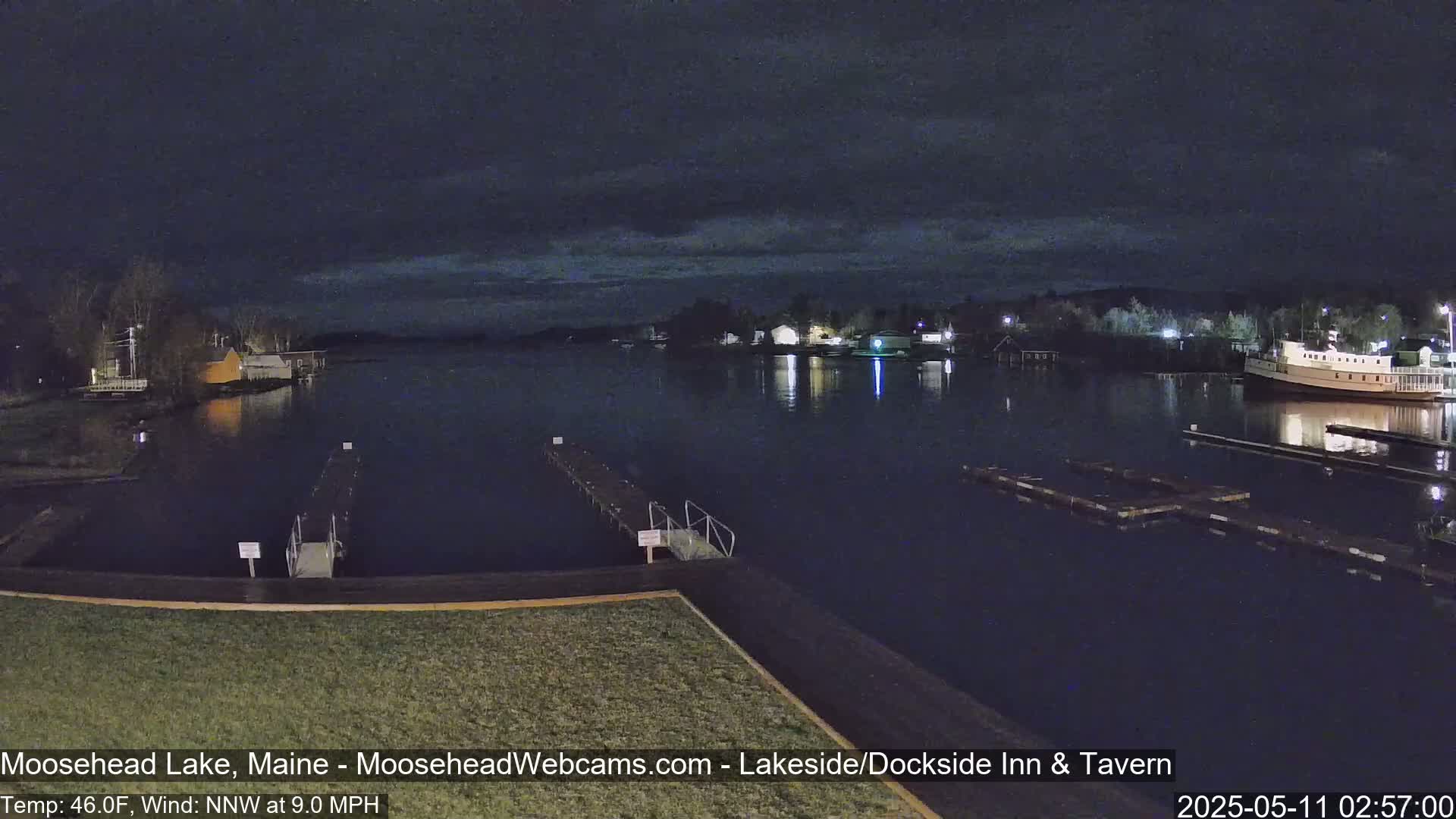 A nighttime view of a calm lake with docks, boats, and buildings illuminated along the shore under a dark, cloudy sky.