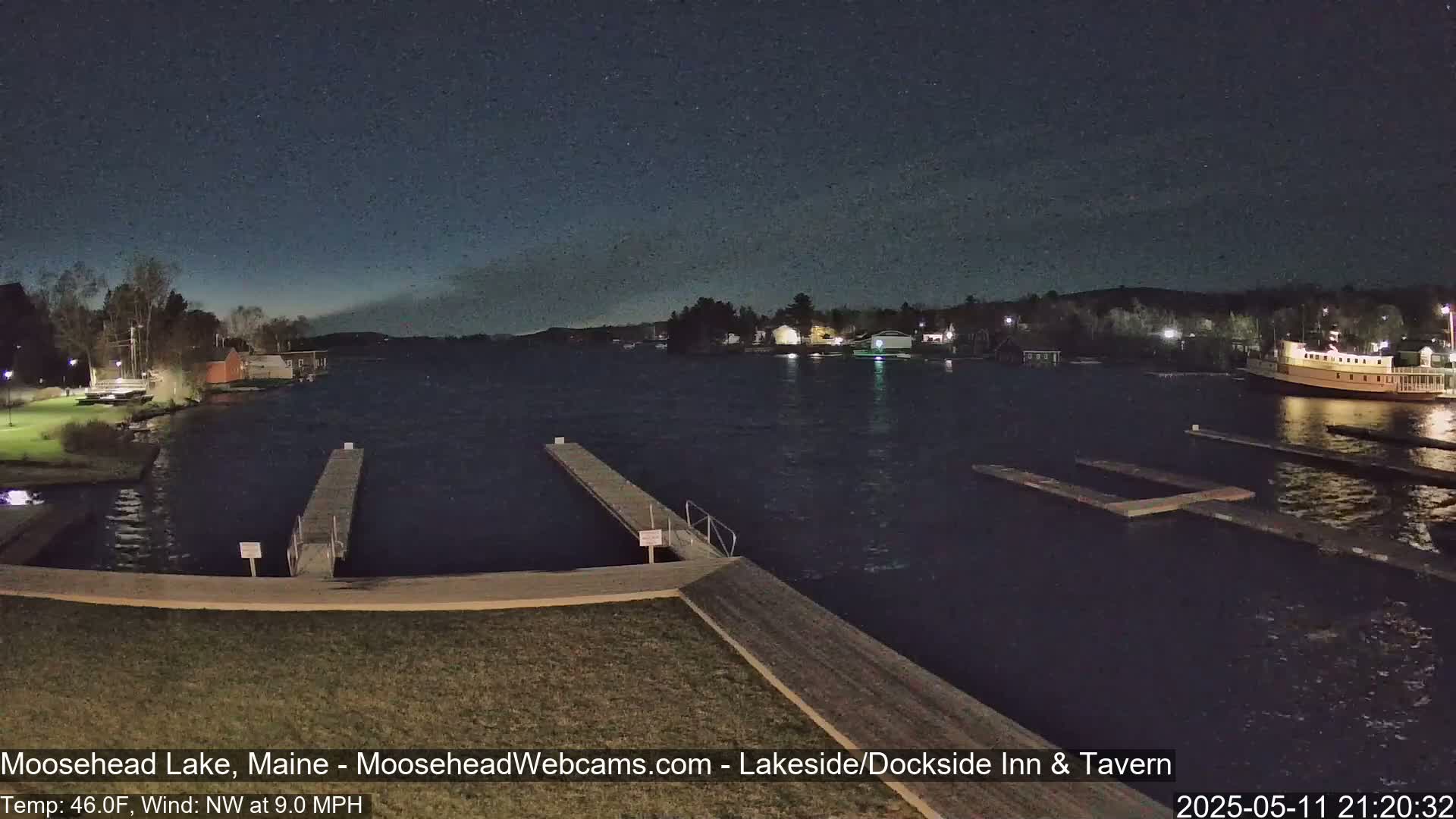 A nighttime view of a lake with docks, a boat, and buildings on the shore under a dark, clear sky.