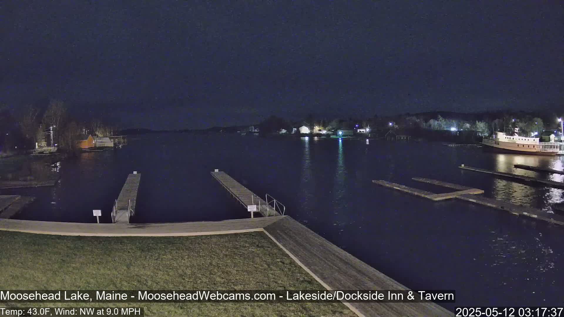 A nighttime view of a calm lake with docks, a boat, and shoreline buildings under a dark sky, with a northwest wind at 9 mph and a temperature of 43 degrees Fahrenheit.