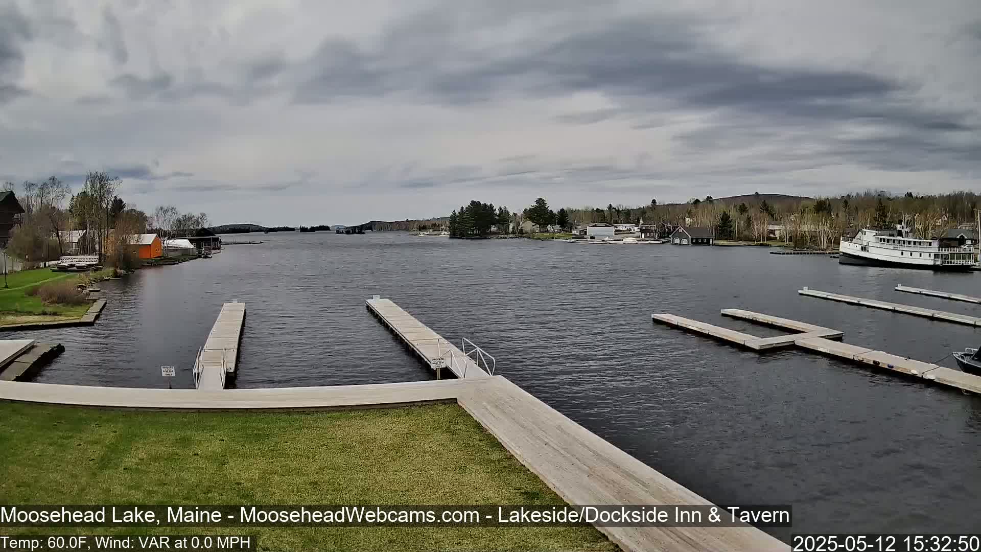 A calm lake with several docks and a boat, surrounded by trees and buildings under a cloudy sky.