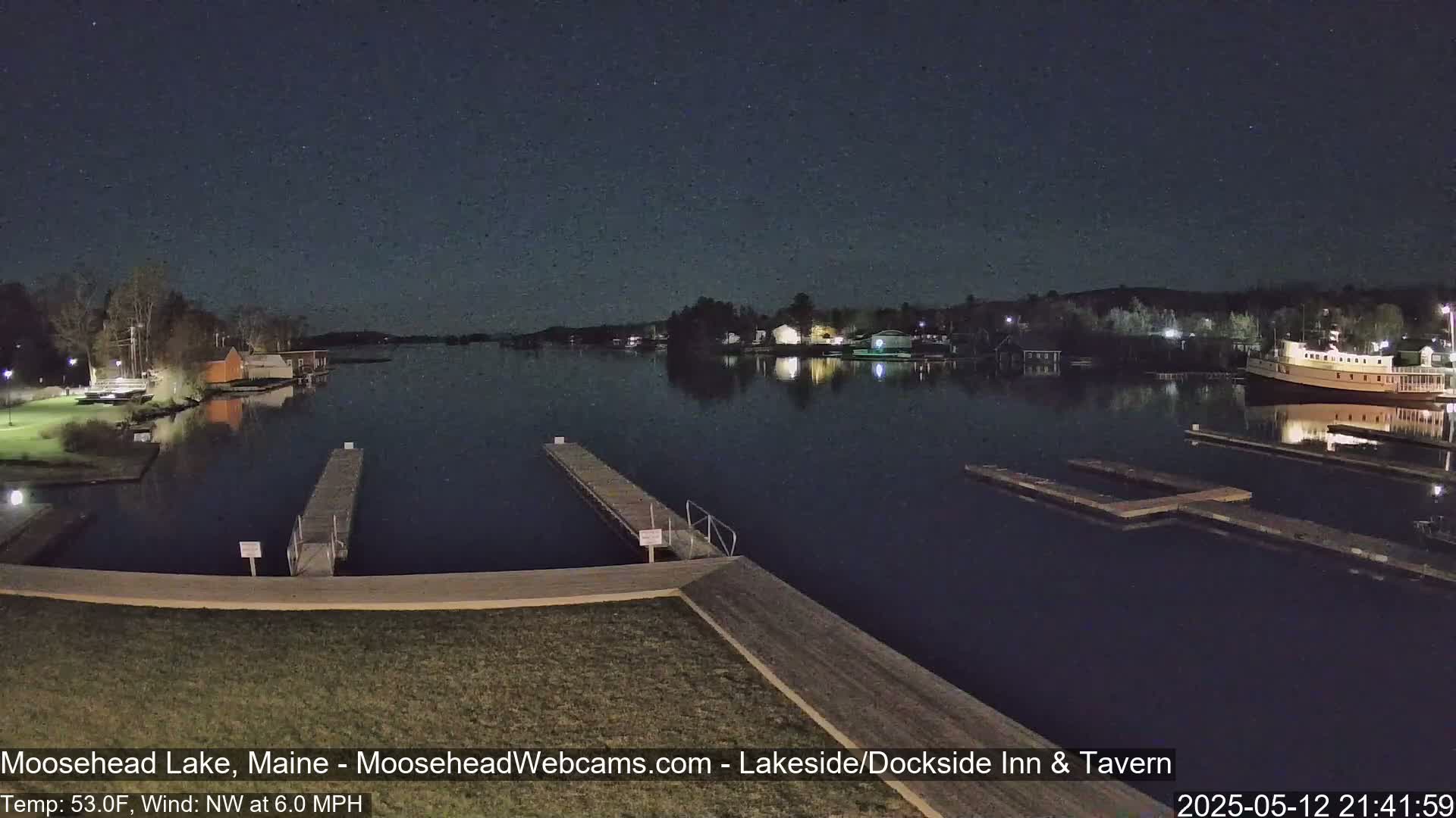 A nighttime view of a calm lake with docks, buildings, and a boat visible under a clear, starry sky.