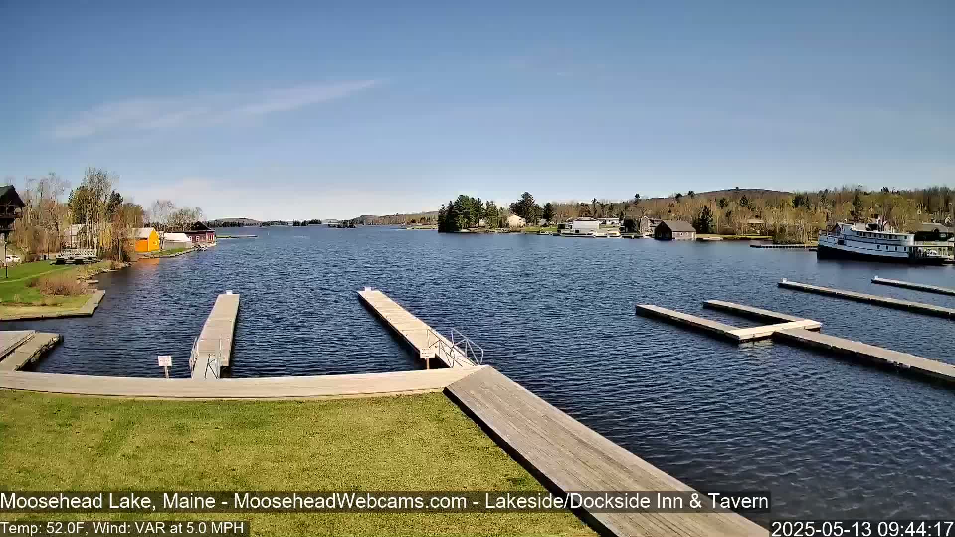 A calm lake with several docks and a boat is seen on a sunny day, with trees and buildings visible on the shore.