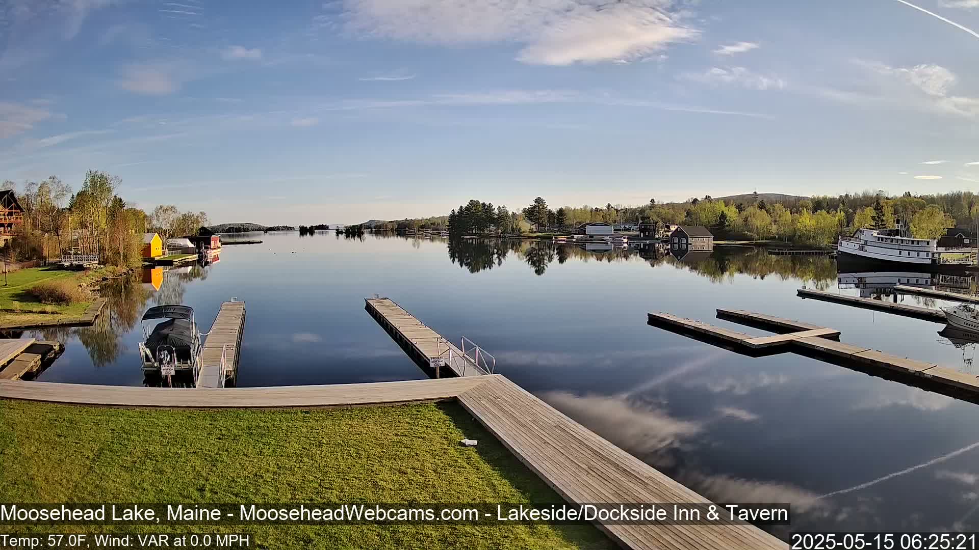A calm lake reflects a clear sky and trees, with boats docked at several wooden piers and a larger vessel moored further out.