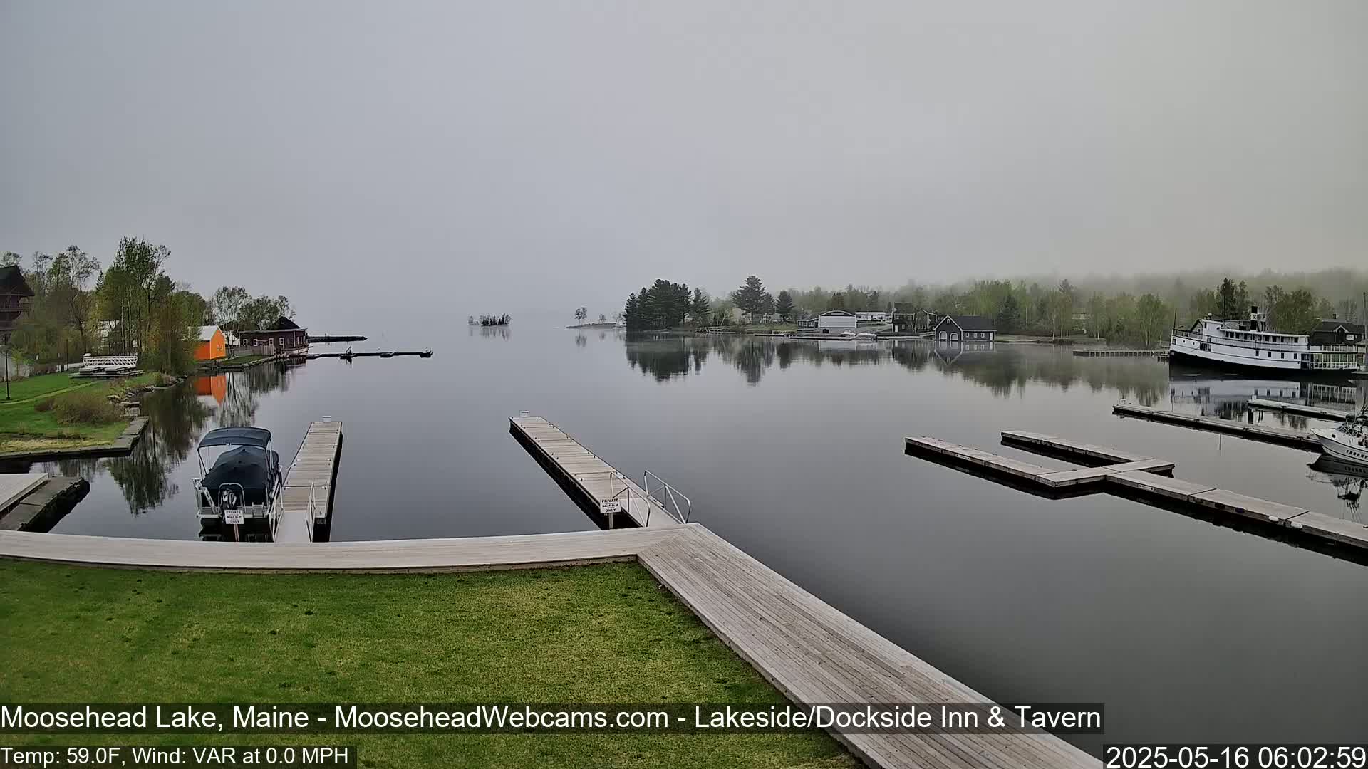 A calm, foggy lake scene shows several docks with boats moored, and buildings on the shoreline under a gray sky.