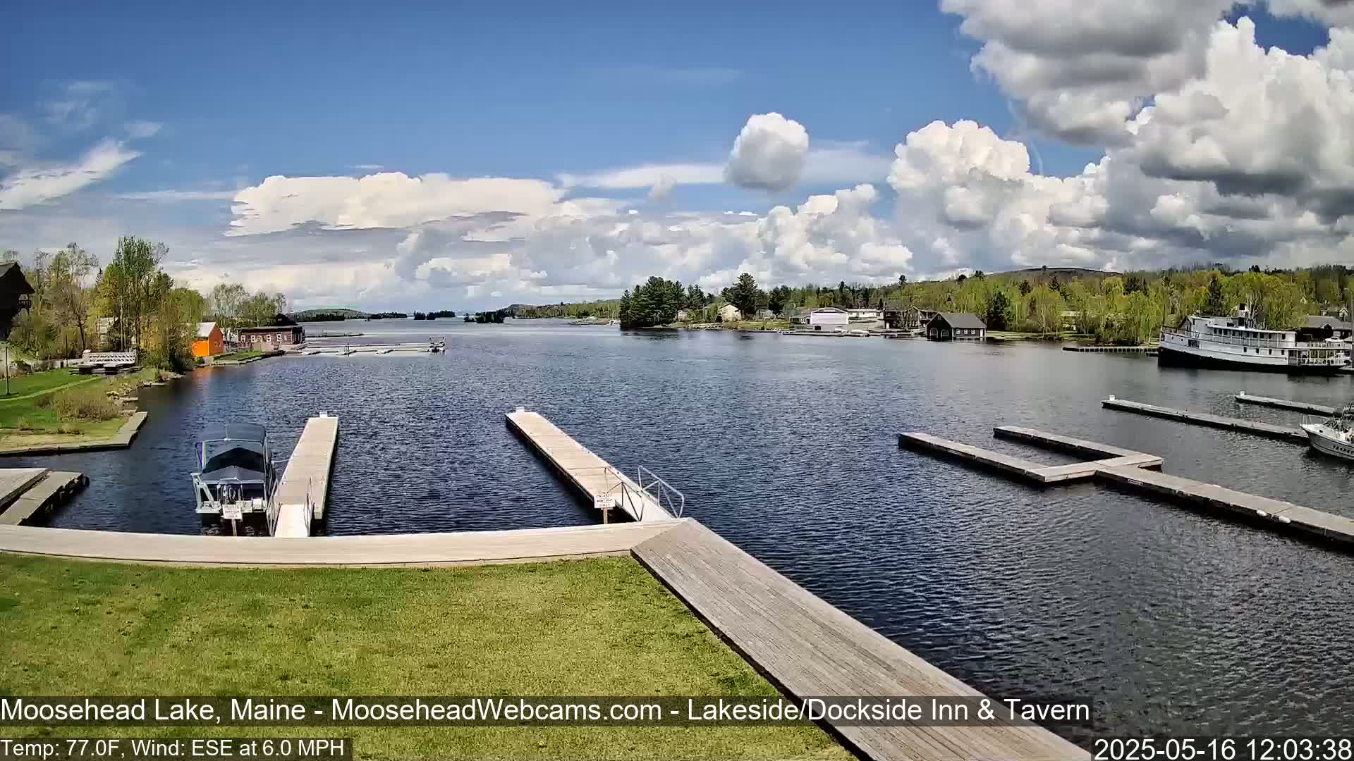 A partly cloudy day shows a calm lake with several docks, boats, and buildings along the shore, lined with green trees.