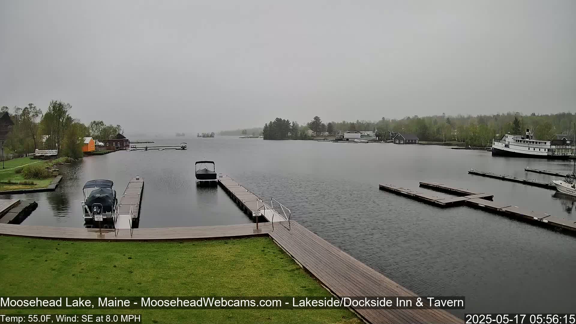 A gray, overcast day on a lake shows several boats moored at docks, with trees and buildings visible across the water.