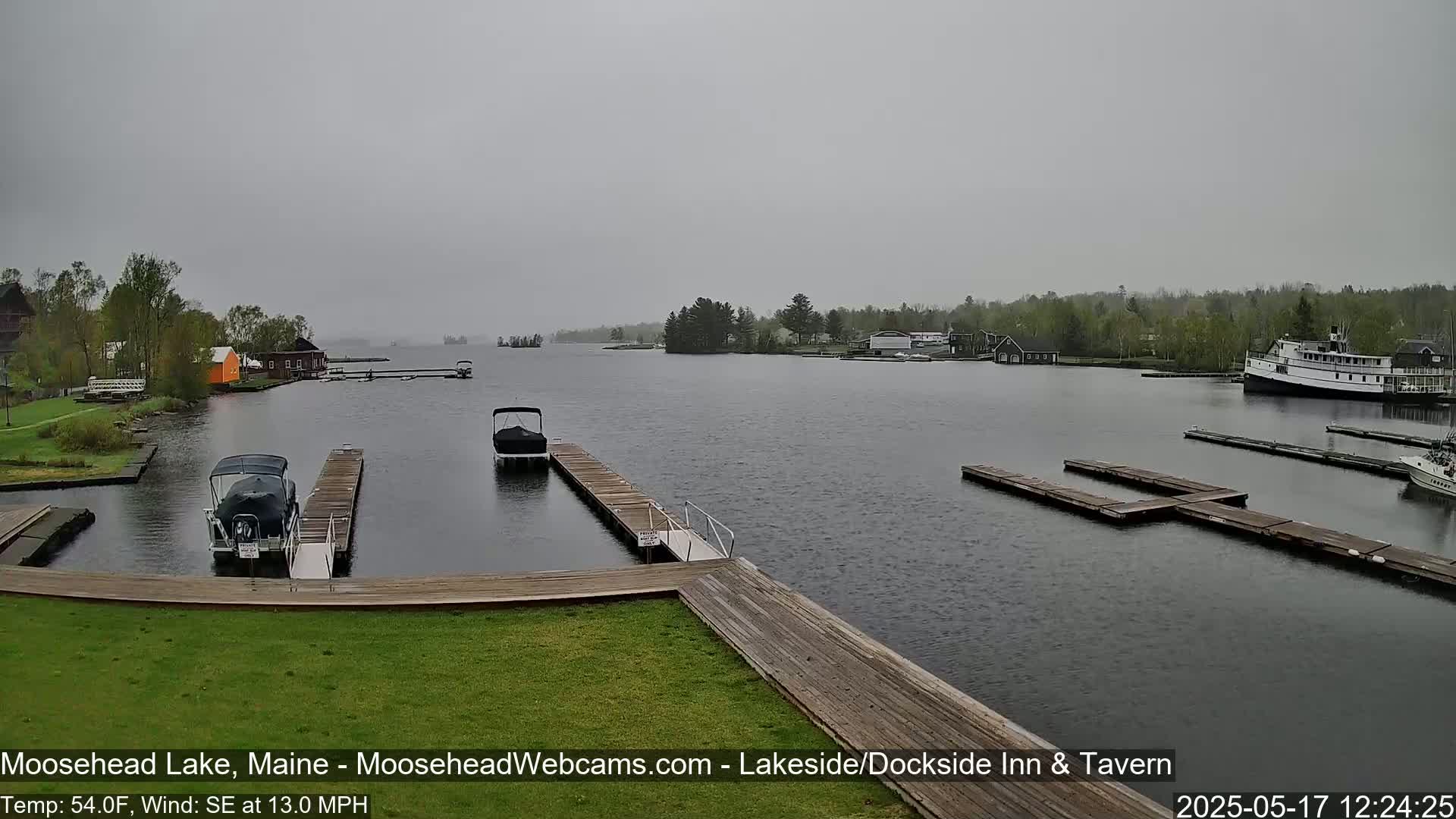 A lake scene on an overcast day shows several boats docked at various piers, with a larger boat visible in the distance.