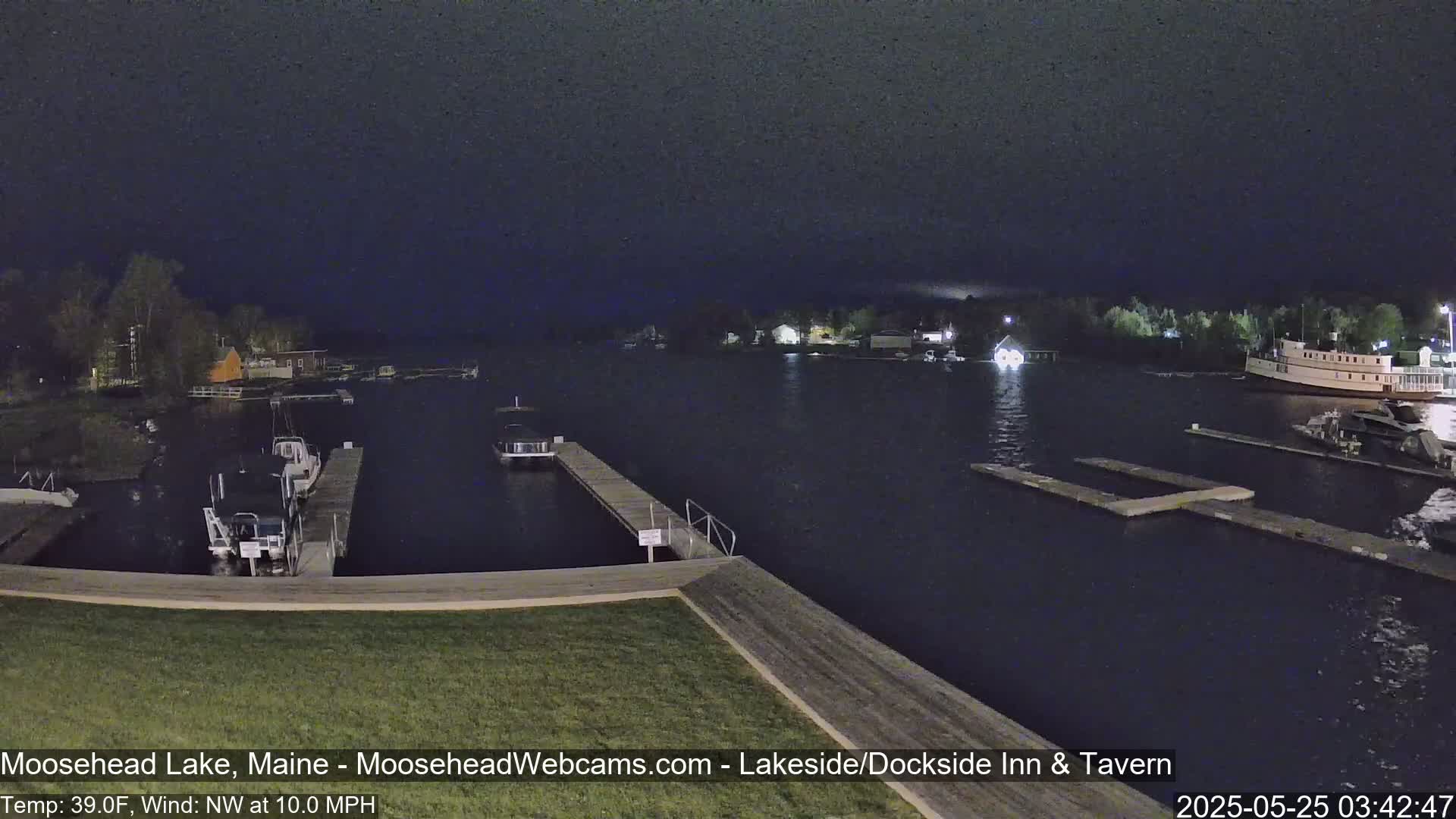 A nighttime view of a calm lake with boats docked at piers under a dark sky, with a northwest wind blowing at 10 mph and a temperature of 39°F.