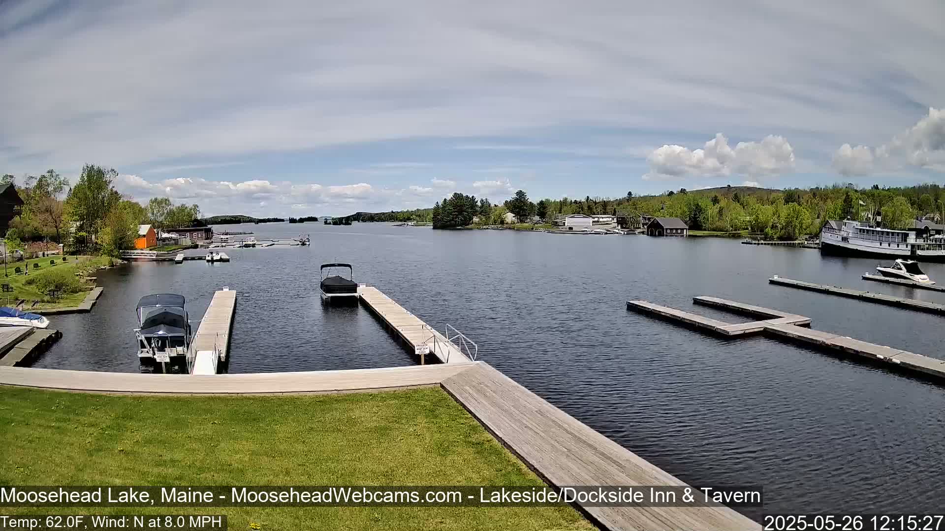 A partly cloudy day on a lake shows several boats docked at various piers, with a larger vessel visible in the distance.