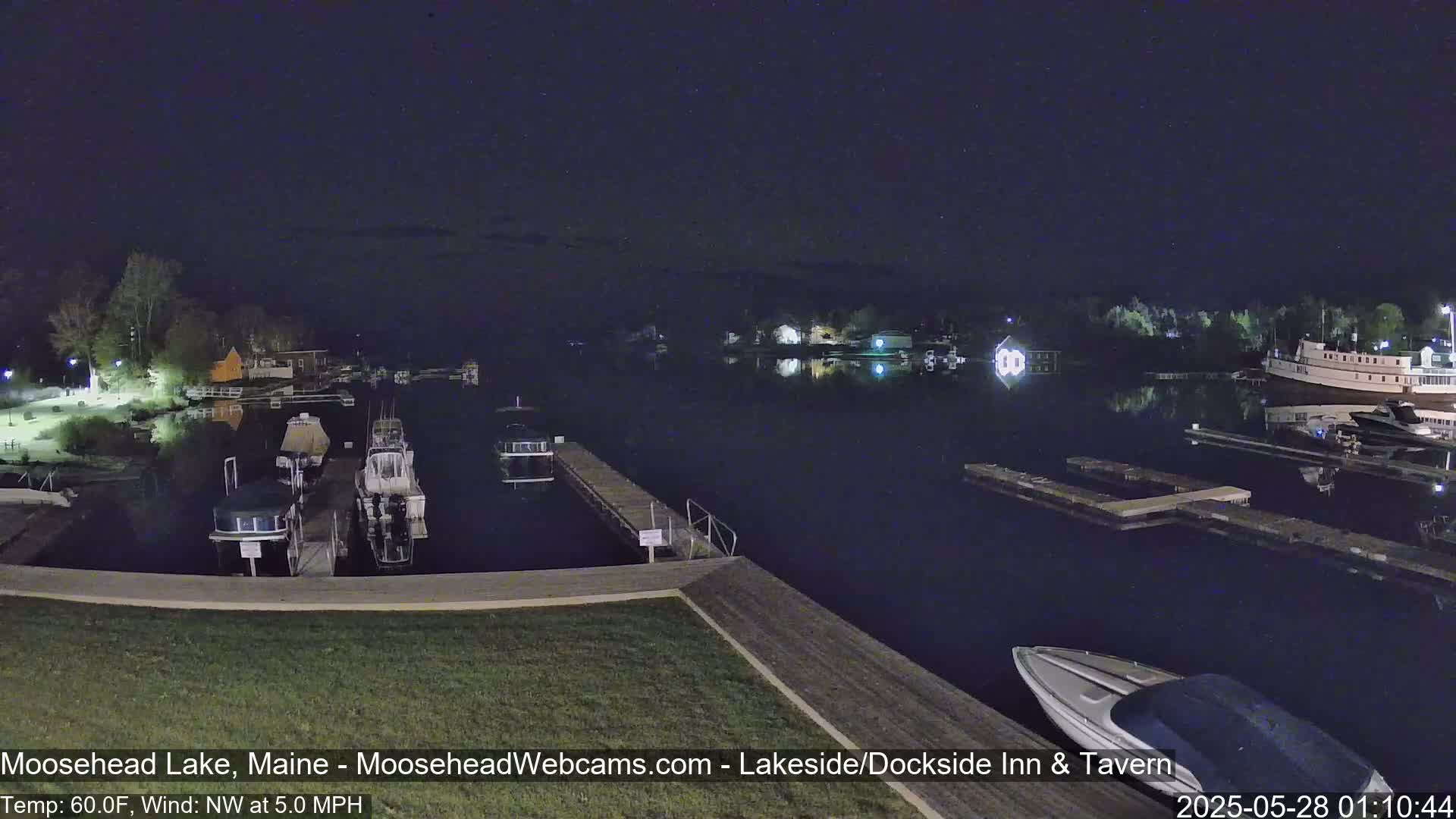 A nighttime view of a calm lake with docked boats and a few lights visible on shore under a dark sky.