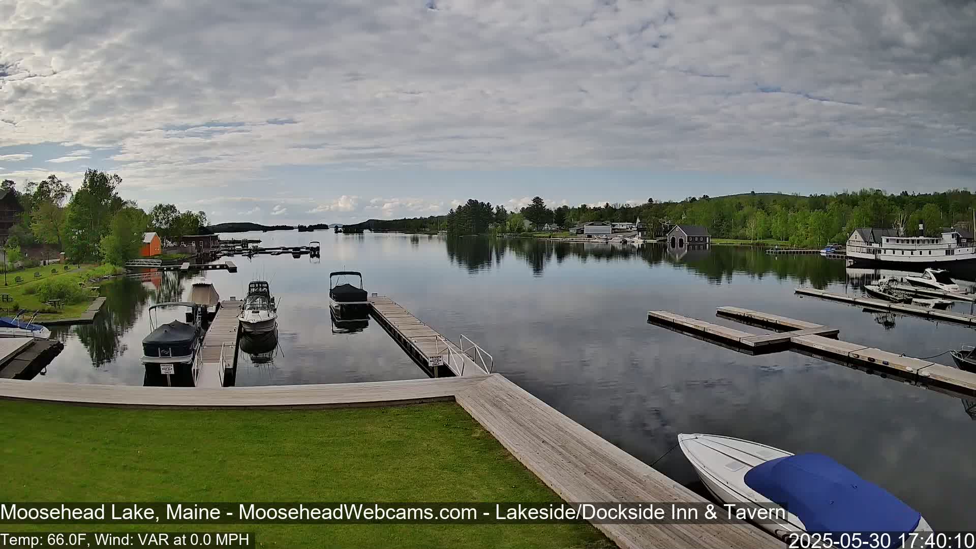 A calm lake with several boats docked at piers, surrounded by green trees and buildings under a partly cloudy sky.
