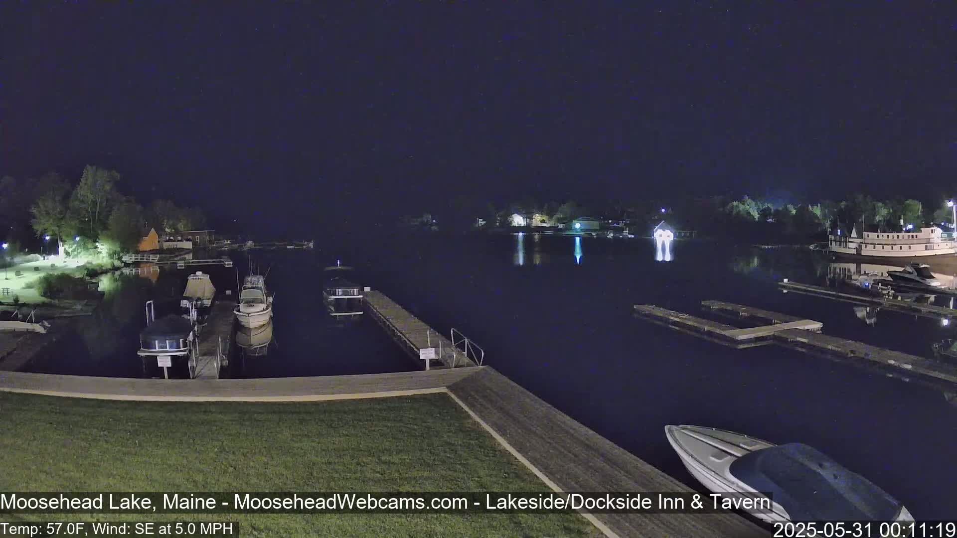 A nighttime view of a calm lake with several boats docked at piers under a dark sky, a gentle southeast wind present.