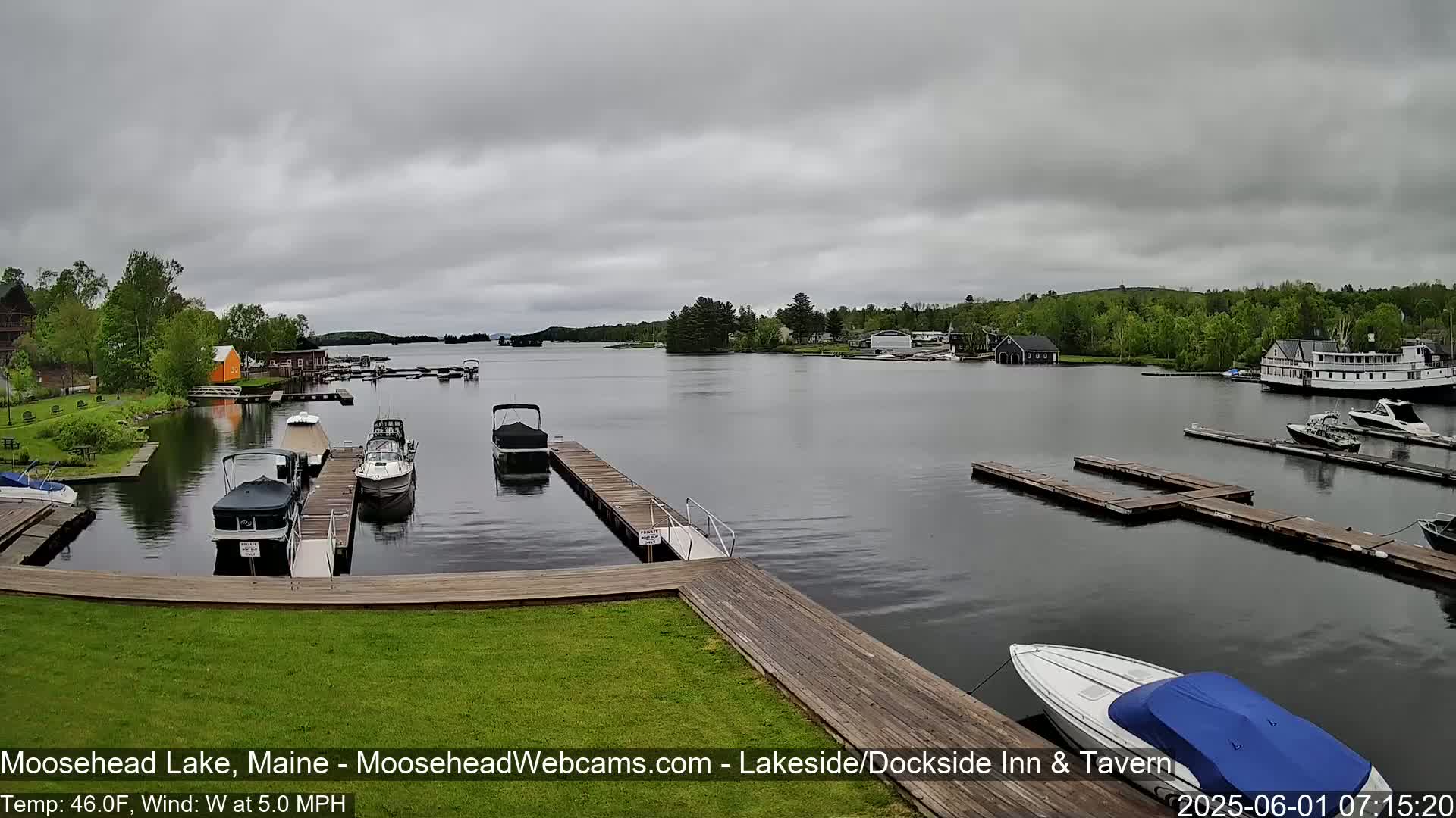 A lake view shows several boats docked at wooden piers under an overcast sky, with a light wind.