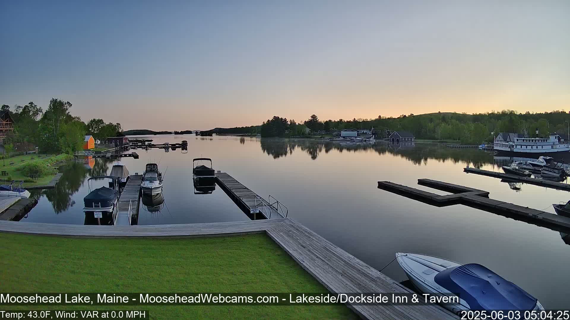Several boats are moored at docks on a calm lake at sunrise, with trees and buildings visible along the shoreline.