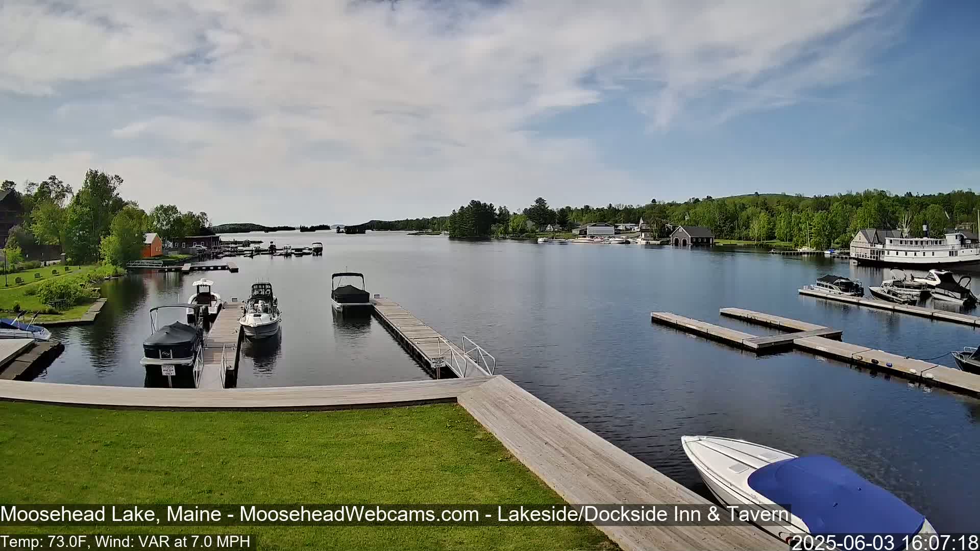A calm lake with numerous boats docked at piers, surrounded by green trees and under a partly cloudy sky.