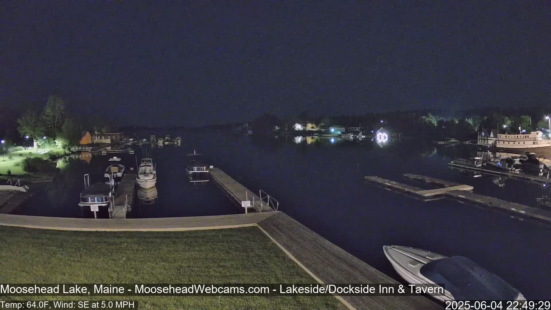 A nighttime view of a calm lake with several boats docked, lit buildings in the distance, and a gentle southeast wind.