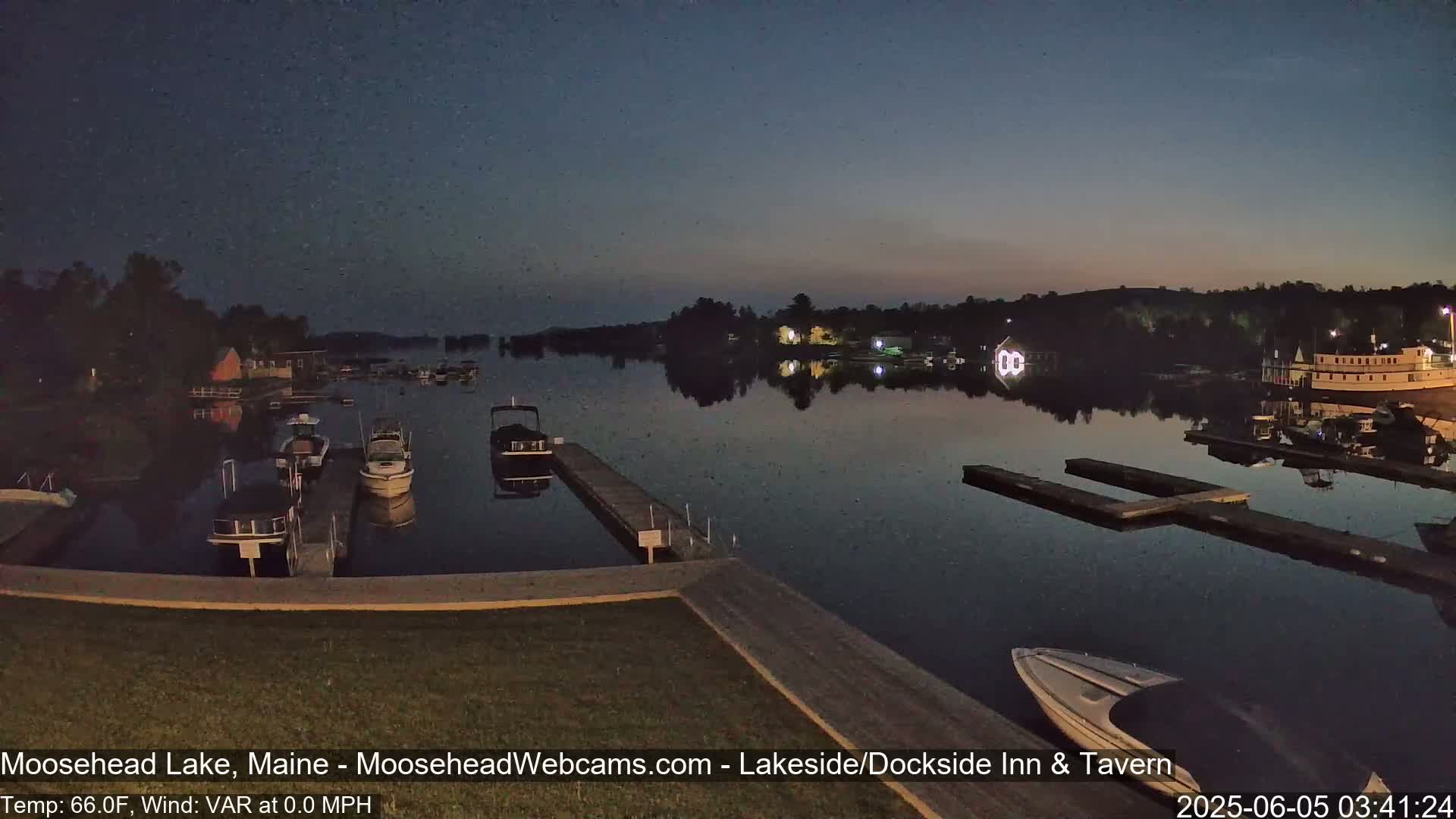 A calm, dark lake at twilight shows several boats docked at piers with buildings and trees visible on the shore.