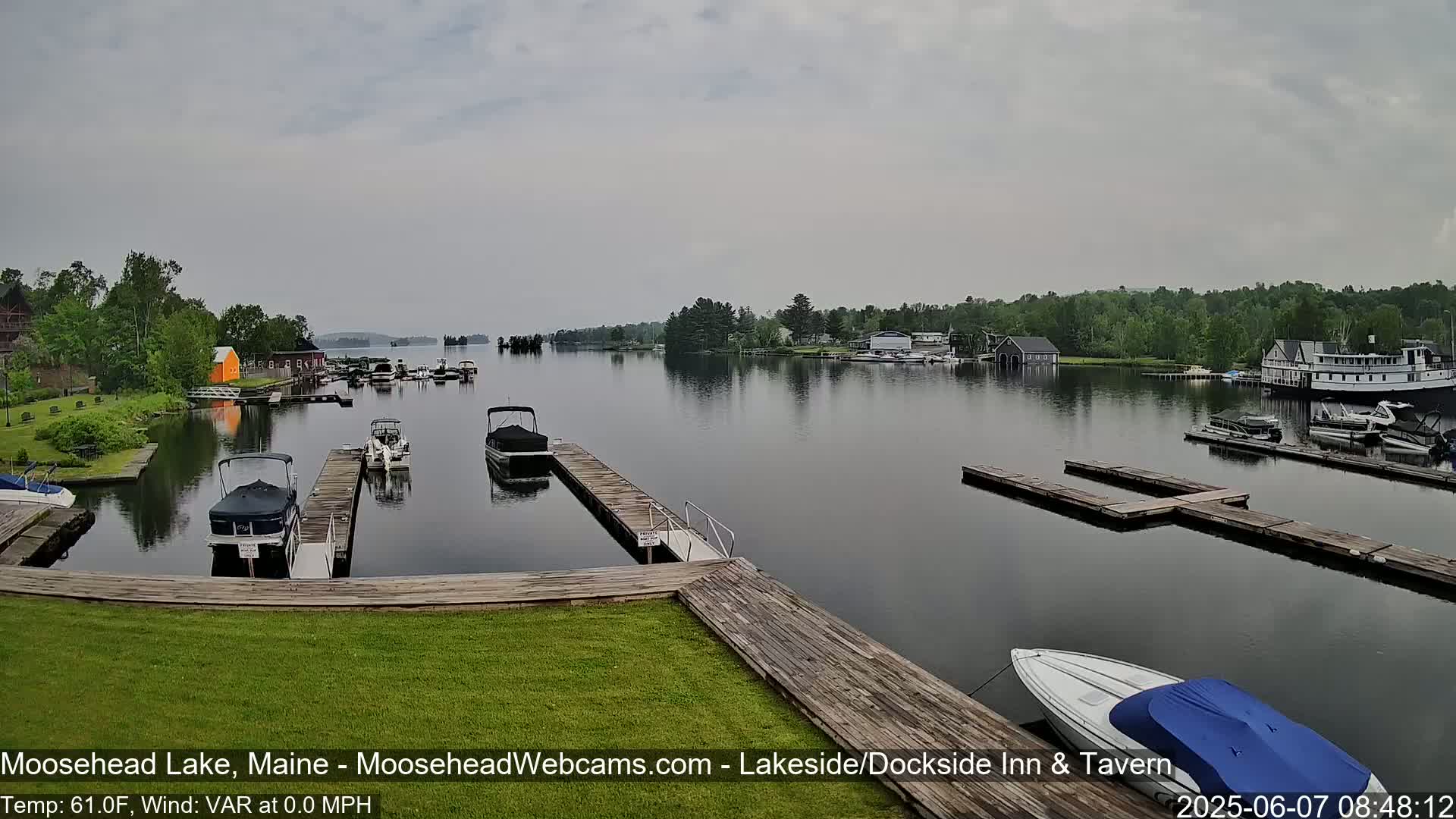 Several boats are docked at a marina on a calm, overcast lake, with trees and buildings visible in the distance.