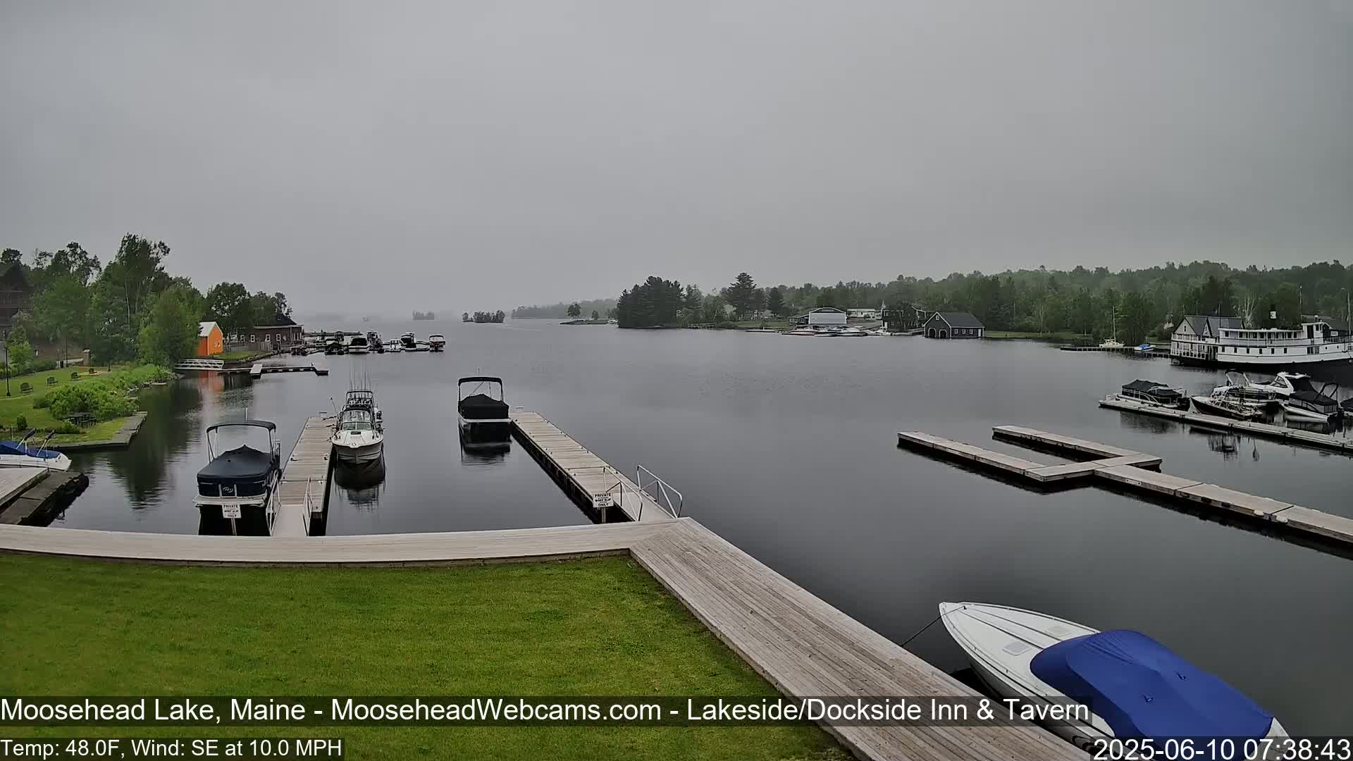 A gray, overcast day shows a calm lake with several boats docked at piers and a few others on the water, surrounded by green vegetation and buildings.