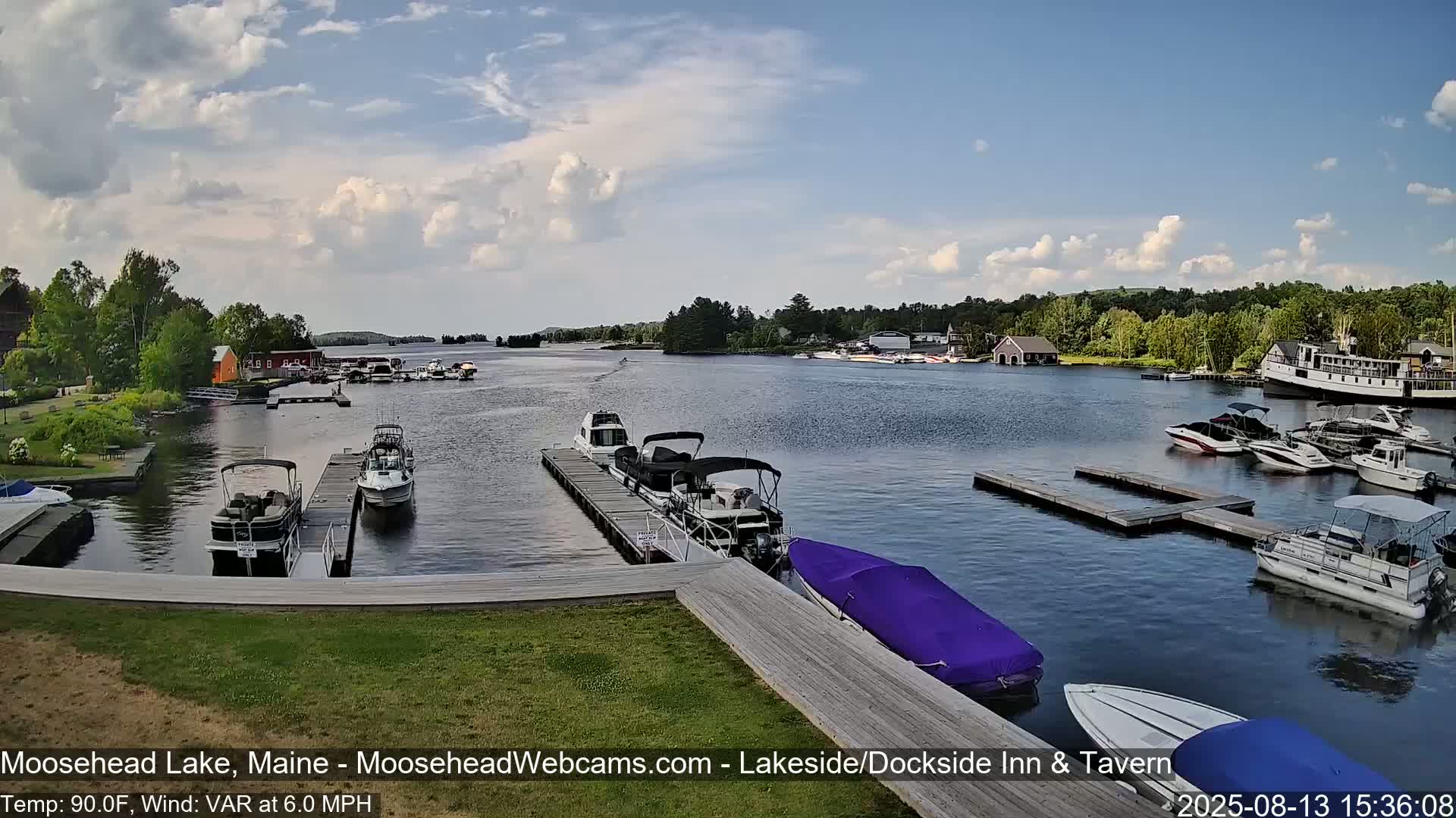 A partly sunny day shows numerous boats docked at a marina on a lake, with several more boats on the water.