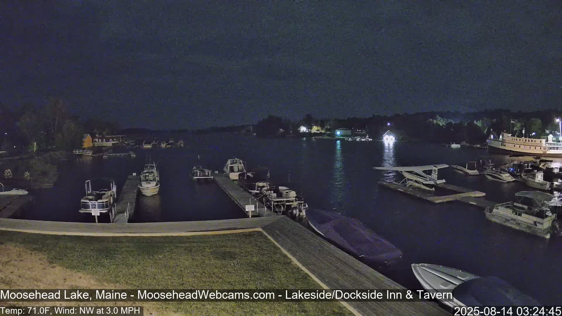 A nighttime view of a calm lake shows several boats docked at piers under a dark sky with a light northwest wind.