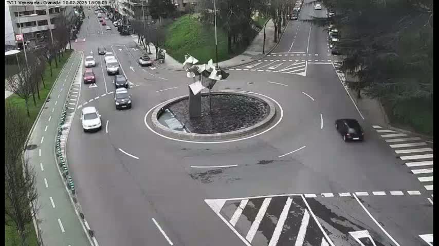 A roundabout with several cars driving around it and on surrounding roads, under a mostly cloudy sky.
