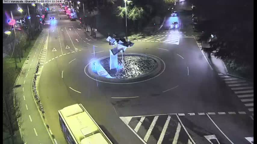 A nighttime aerial view shows a roundabout with a modern sculpture in the center, surrounded by roads with sparse traffic.