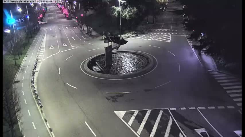 A nighttime aerial view shows a mostly empty roundabout with a dark sculpture in the center, surrounded by roads with white lane markings and a crosswalk.