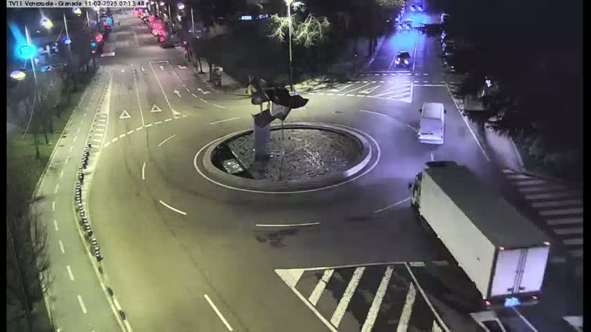 A nighttime aerial view shows a roundabout with a sculpture in the center, several vehicles navigating the roads, and a large truck approaching the roundabout.