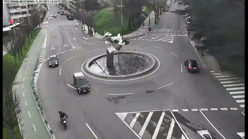 A traffic roundabout with a modern sculpture fountain in the center, surrounded by several cars and a motorcycle, on a mostly overcast day.