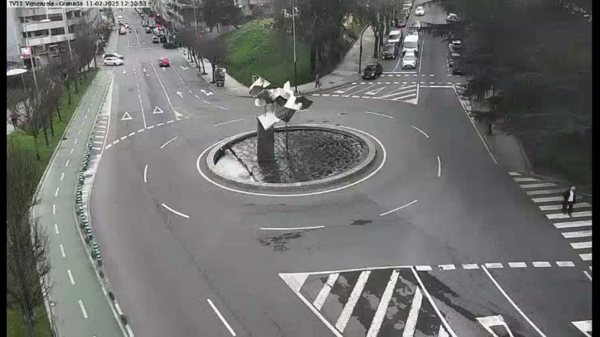A roundabout with a modern abstract sculpture in its center is surrounded by roads with sparse traffic, under an overcast sky.